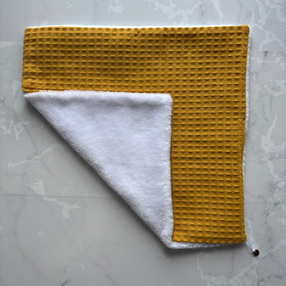 serviette visage posé à plat en éponge bambou blanc et nid d'abeille moutarde avec un logo médaillon.
