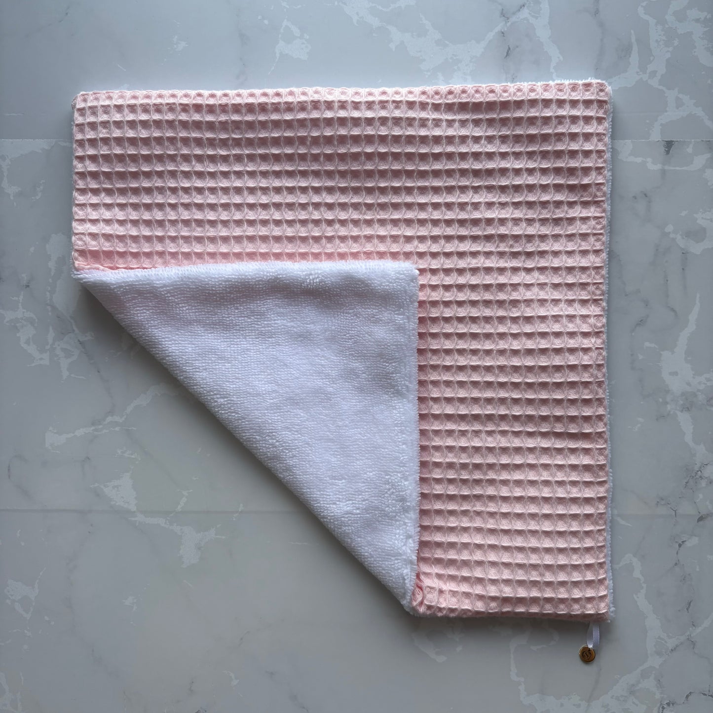 serviette visage posé à plat en éponge bambou blanc et nid d'abeille rose pale avec un logo médaillon.
