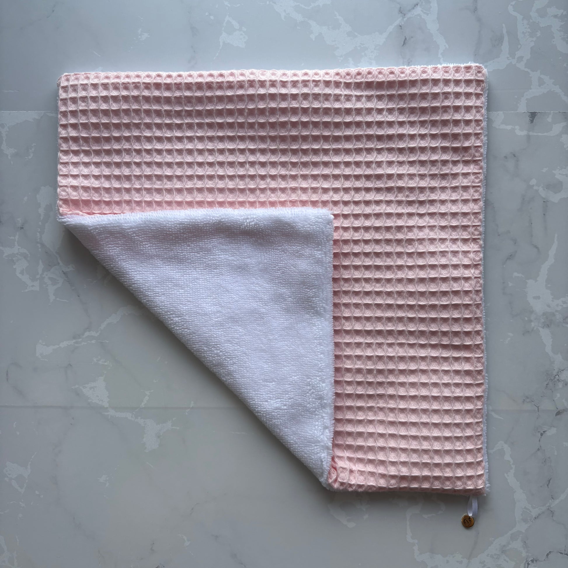 serviette visage posé à plat en éponge bambou blanc et nid d'abeille rose pale avec un logo médaillon.